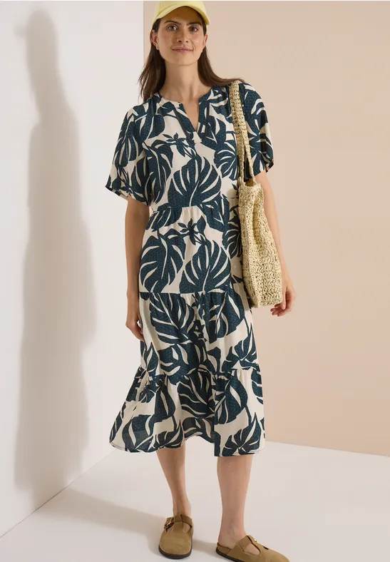 Frau trägt midi Kleid mit navy-blauem Blattmuster, kurzen Ärmeln, Strohtasche und beigen Slippern vor neutralem Hintergrund.