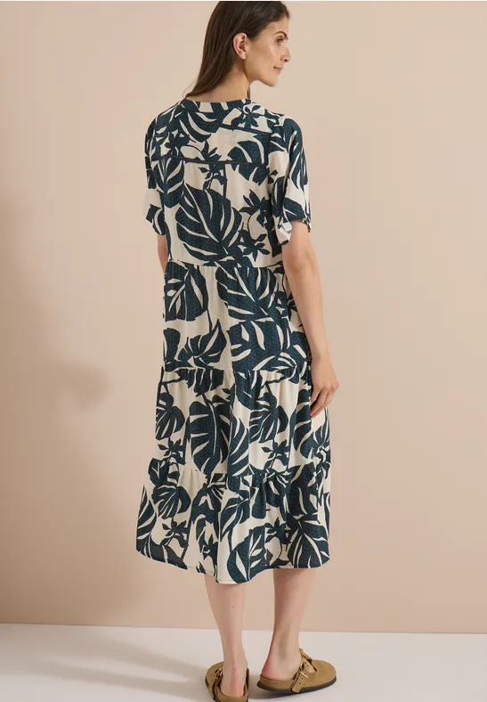 Frau von hinten in midi Kleid mit blauem Blattprint auf beigem Grund und Sandalen.
