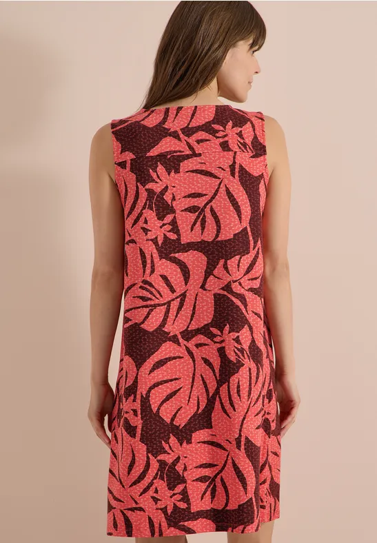 Frau von hinten in ärmellosem roten Kleid mit großem rosa Blattmuster vor beigem Hintergrund.