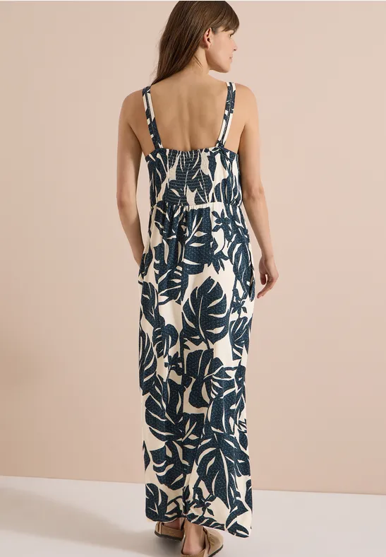 Frau von hinten in ärmellosem Maxikleid mit blauem Blattmuster vor hellem Hintergrund.