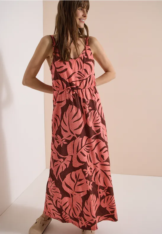 Frau in rotem Maxikleid mit pinkem Blättermuster, steht lächelnd mit Händen in der Hüfte.