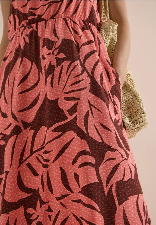 Rotes Maxikleid mit tropischem Blattprint, Frau mit Händen in den Taschen und Basttasche am Arm.