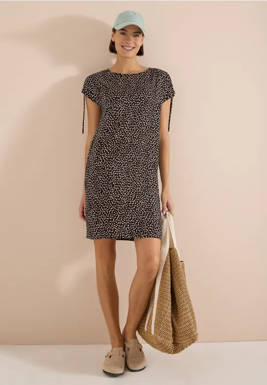 Femme en robe courte noire et blanche à pois, chapeau, mules et sac en paille sur fond beige.