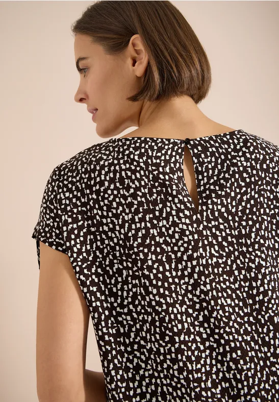 Vue de dos d'une femme aux cheveux courts bruns portant une blouse noire à petits motifs blancs.