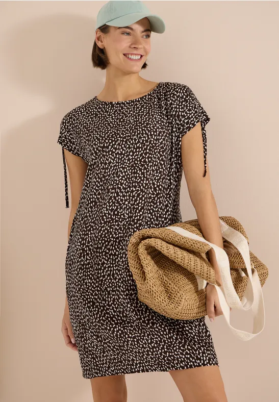Femme en robe à manches courtes à motifs, portant un sac de plage en paille et une casquette.