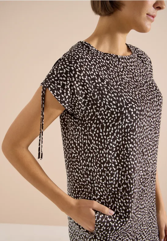 Femme portant un haut noir à motifs blancs, manches courtes avec lien latéral, main dans la poche.