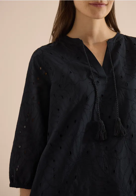 Vrouw draagt zwart geborduurd blouse met oogjes, V-hals en kwastjes aan de koordsluiting.