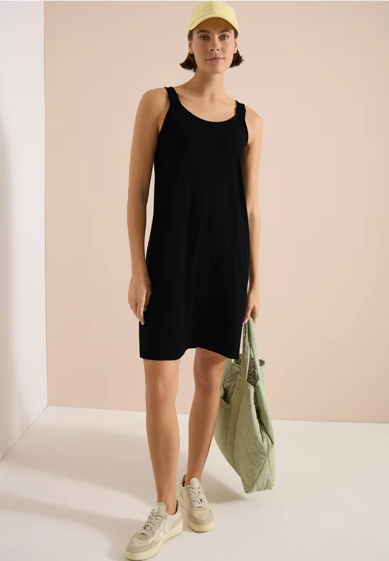 Schwarzes ärmelloses Kleid, helle Sneaker, grün-gestreifte Tasche, schlichter Raum.