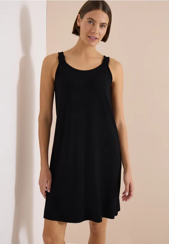 Frau in schlichtem schwarzen, ärmellosem Kleid vor hellem Hintergrund.