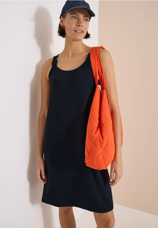 Person trägt ärmelloses schwarzes Kleid, orange gesteppte Tasche und Baseballcap vor neutralem Hintergrund.