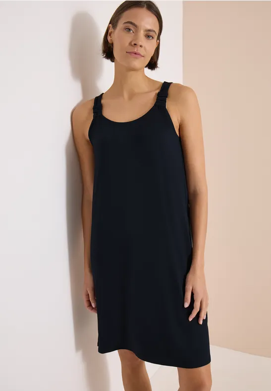 Eine Frau in einem schwarzen ärmellosen Kleid lehnt an einer Wand.