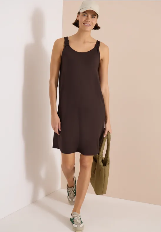 Frau in ärmellosem dunklen Kleid und Sneakers vor beiger Wand, hält grüne Tasche.