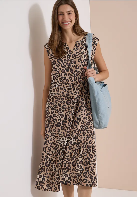 Eine Frau in langem Leopardenkleid mit hellblauer Tasche steht vor einer neutralen Wand.