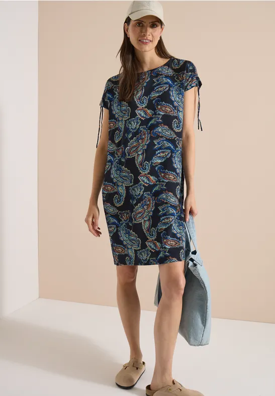 Femme en robe courte imprimé cachemire à col V, casquette, sandales, tenant une veste en jean.