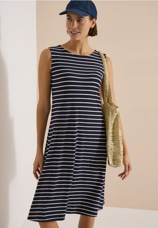 Femme en robe midi rayée bleu et blanc, sac en paille et casquette devant un mur clair.