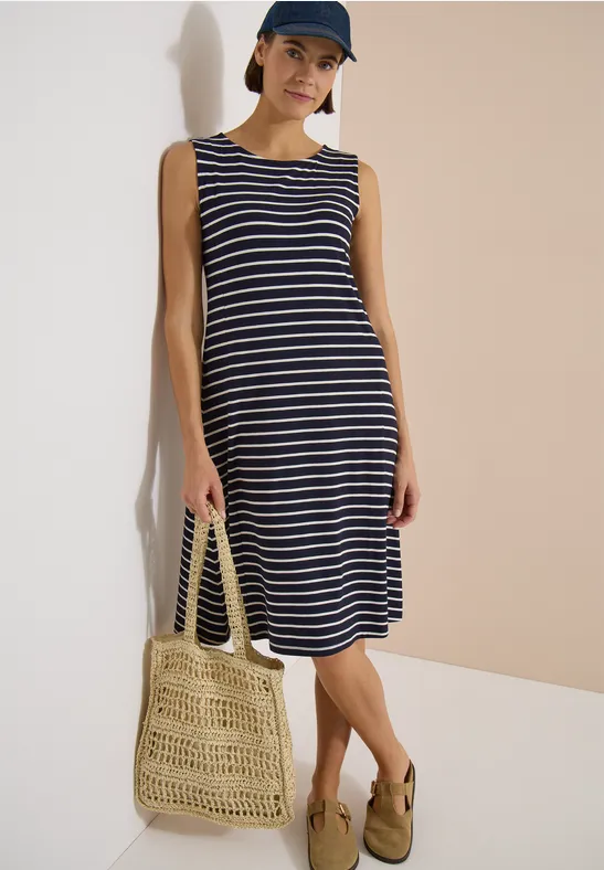 Femme en robe sans manches rayée bleu-blanc, sac en paille tissé et sandales, fond neutre.