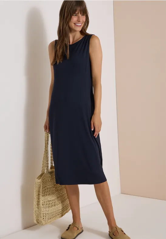 Vrouw in donkerblauwe mouwloze midijurk, strozak en groene espadrilles.