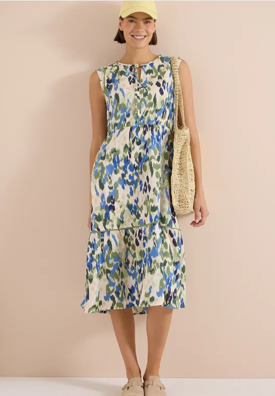 Frau im ärmellosen Midikleid mit blau-grünem Blumenmuster und Strohtasche vor beiger Wand.