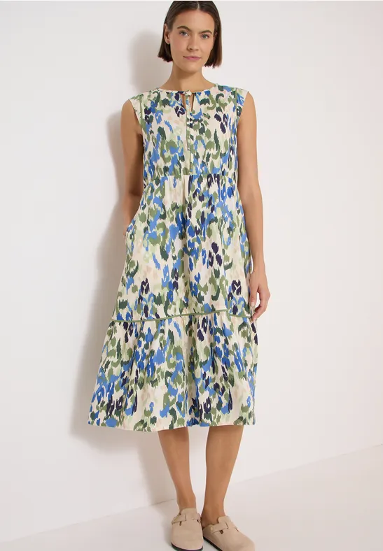 Frau steht vor weißem Hintergrund, trägt ärmelloses Midikleid mit grün-blauem Blumenmuster und Stufenrock.