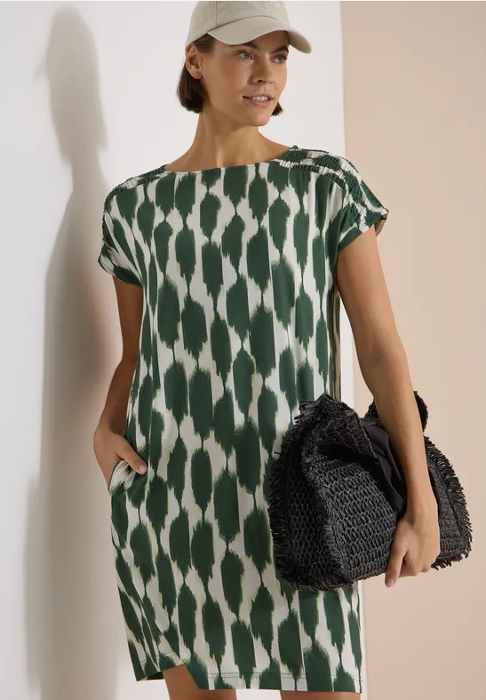 Dame trägt ein locker sitzendes grün-weiß gemustertes Kleid mit kurzen Ärmeln und hält eine schwarze Flecht-Handtasche.
