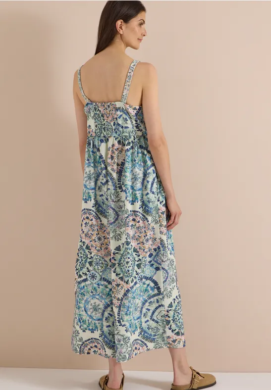 Femme de dos portant une robe longue imprimée à fines bretelles, fond beige neutre.