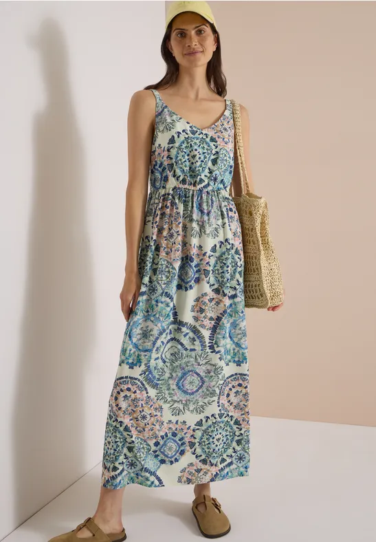 Femme en robe longue sans manches à motifs mosaïque bleu-vert, sac en paille et sandales plates.