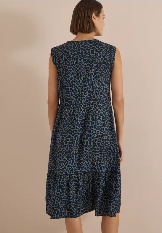 Rugaanzicht: vrouw in mouwloze midi-jurk met blauw-zwart dierenprint, voor een effen beige achtergrond.