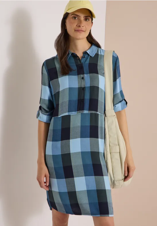 Femme en robe-chemise à carreaux bleus, casquette et sac beige, devant un fond neutre.