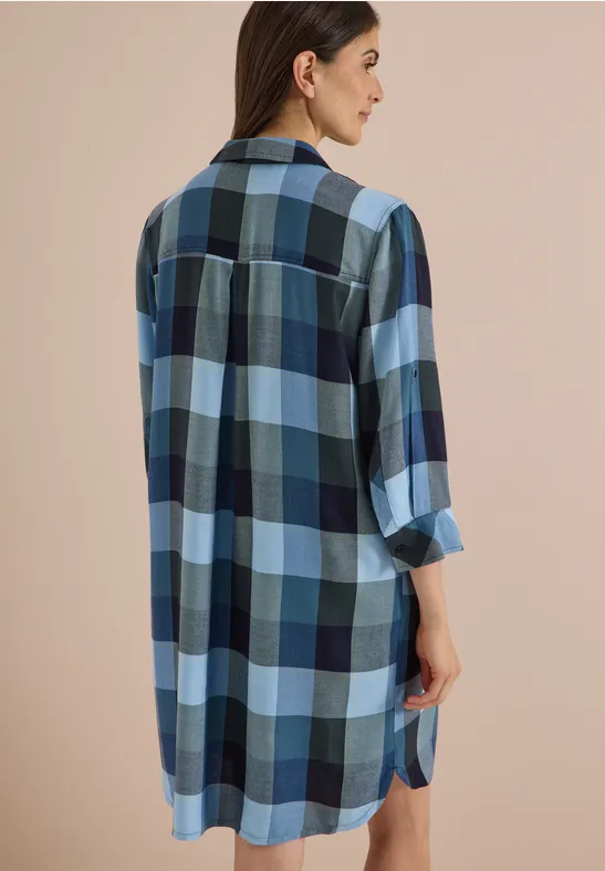 Femme de profil/dos, portant une chemise-robe ample à carreaux bleus, blancs et noirs, manches longues.