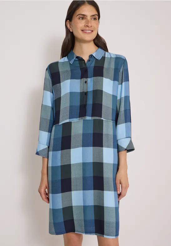 Une femme porte une robe-chemise à carreaux bleus aux manches 3/4.