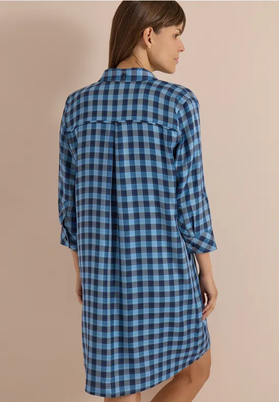 Frau von hinten in blau kariertem Hemdkleid vor beigem Hintergrund.