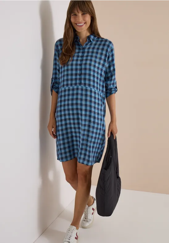 Lächelnde Frau in blau-kariertem Hemdkleid, Sneakers und Tasche.