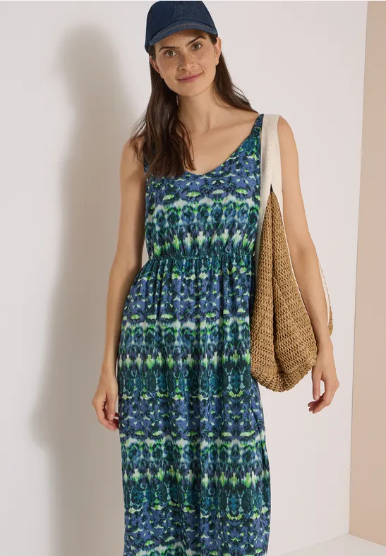Femme en robe longue sans manches bleu-vert à motifs, casquette et sac tressé, fond neutre.