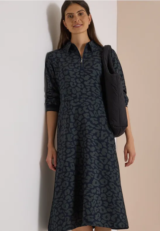 Frau in Midi-Hemdkleid mit grün-blauem Muster und Schulter-Tasche vor hellem Hintergrund.