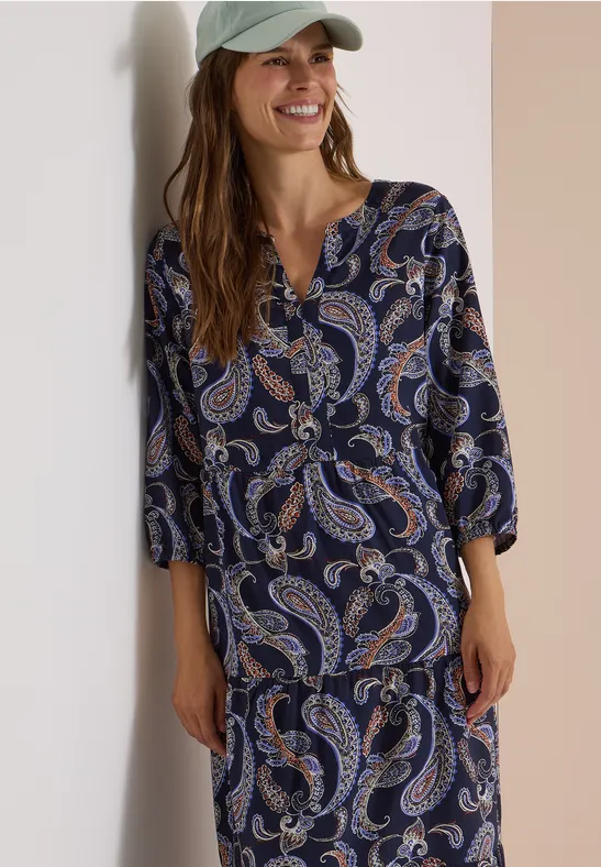 Frau in blauem Paisley-Oberteil und passendem Rock lächelt.