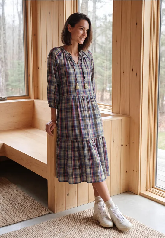 Vrouw in geruite jurk en witte cowboylaarzen staat in een lichte houten ruimte bij het raam.