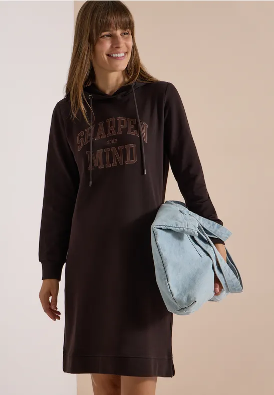 Vrouw lacht, draagt een bruin hoodie-jurk met opdruk en houdt een lichtblauwe tas vast.