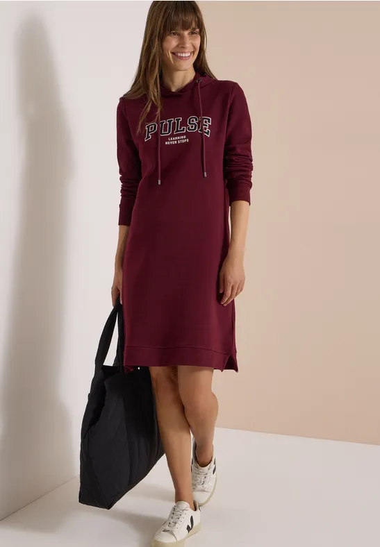 Frau in burgunderfarbenem Kapuzenkleid mit "Poler"-Schriftzug, hält schwarze Tasche und trägt weiße Sneaker.