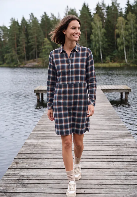 Eine Frau in kariertem Hemdblusenkleid spaziert lächelnd auf einem Holzsteg am ruhigen See, umgeben von Wald.