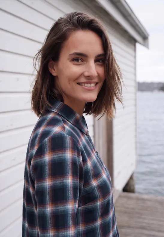 Eine lächelnde Frau in karierter Bluse lehnt an einer weißen Holzwand am Wasserpier.