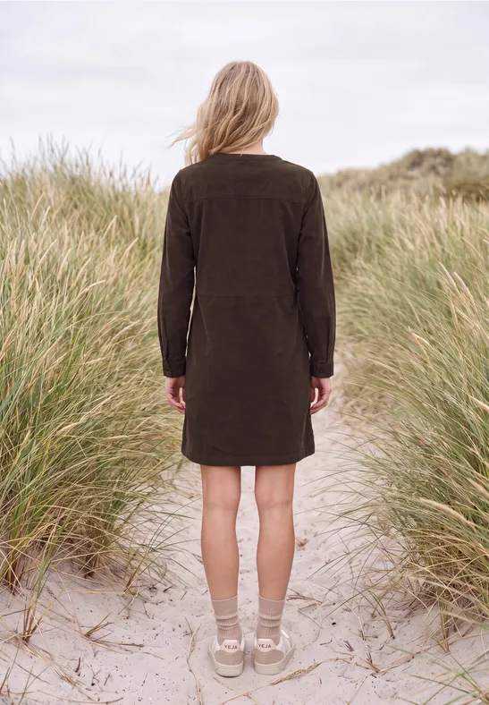 Une femme en robe marron, de dos, se tient sur des dunes de sable entourées d’herbes de plage.