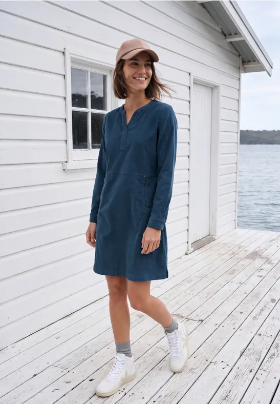 Frau in blauem Langarmkleid steht lächelnd auf einem Holzsteg vor einem weißen Haus am Wasser.