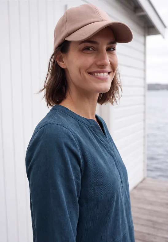 Lächelnde Frau mit Baseballcap und blauem Oberteil steht am Wasser vor einem weißen Gebäude.