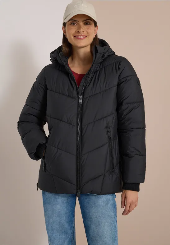 Frau mit schwarzer Steppjacke, blauer Jeans und weißer Cap lächelnd vor hellem Hintergrund.