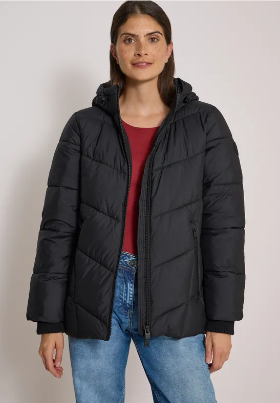 Eine Frau in einer schwarzen Steppjacke mit Kapuze, rotem Oberteil und Jeans vor hellem Hintergrund.