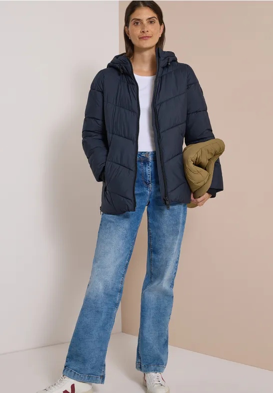 Frau in dunkelblauer Steppjacke, weißem Shirt, blauen Jeans, hält eine olivgrüne Tasche vor neutralem Hintergrund.
