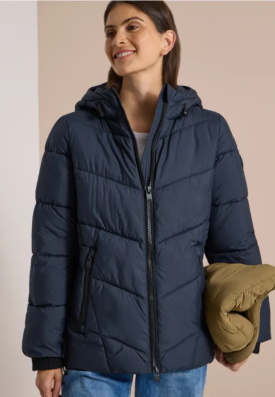 Frau trägt dunkelblaue Steppjacke mit Kapuze, lächelt und hält eine gefaltete khakifarbene Jacke.