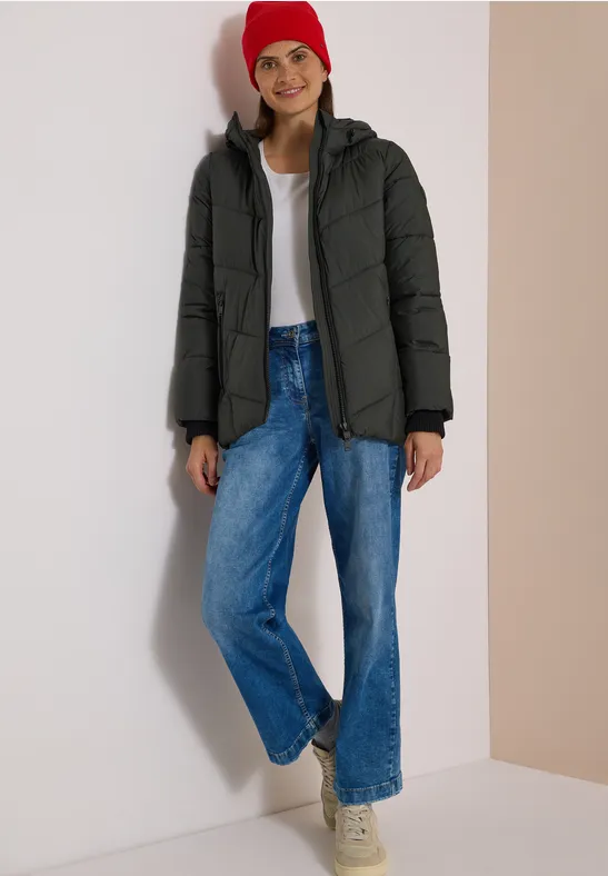 Eine Frau mit roter Mütze, schwarzer Daunenjacke, weißem Shirt, blauen Jeans und Sneakern.