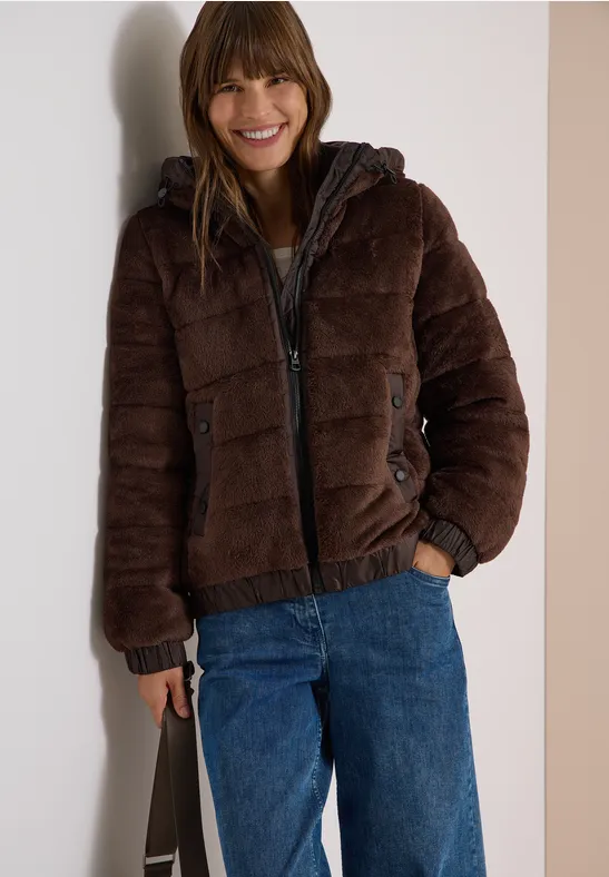 Junge Frau lacht, trägt braune Steppjacke, blaue Jeans und hält eine Tasche.