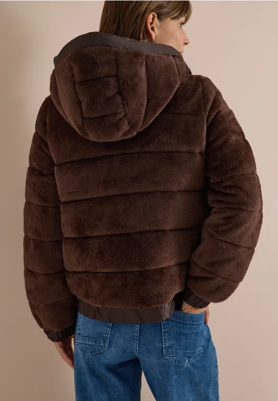 Person von hinten in brauner, gepolsterter Kapuzenjacke und blauen Jeans vor neutralem Hintergrund.